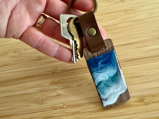 Walnut wood keychain with leather trap and gold ring, decorated with resin ocean waves in blue, turquoise, and white foam.