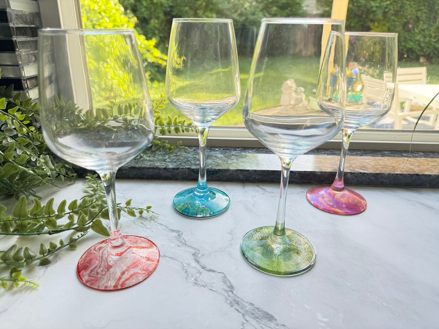 Set of four unique wine glasses. The base of the glasses are decorated with different colors using acrylic paint and sealing them with food safe resin. There is a red and white glass, blue and green, pink and purple, and green and gold.