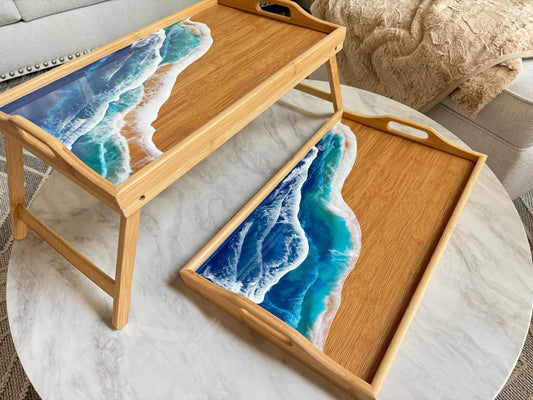 Two Breakfast Trays are pictured. One is folded flat and the other is tall with the legs unfolded. They are decorated with resin ocean waves in bright blue, turquoise, and white. The waves are in the diagonal and cover about half the inside of the tray. the trays are a medium light bamboo color.