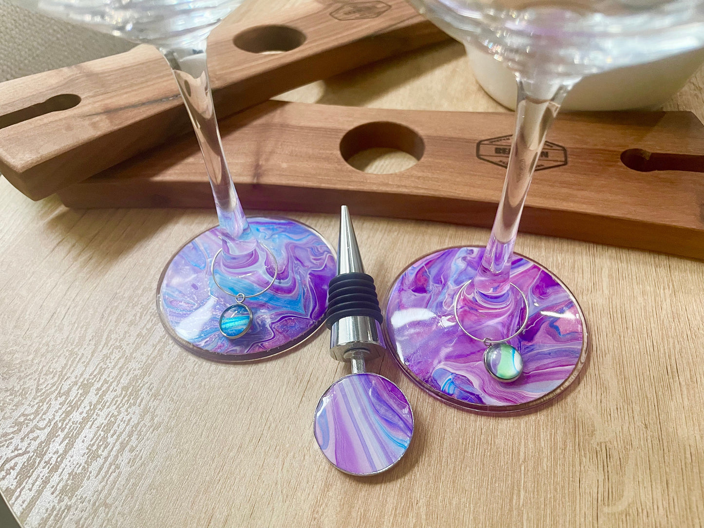 Two hand painted wine glasses in purple, blue, and pink swirls with matching wine bottle stopper, and two wine glass charms. The stopper is double sided and the charms contrast a bit with the painted glass bases to make your glass stand out.